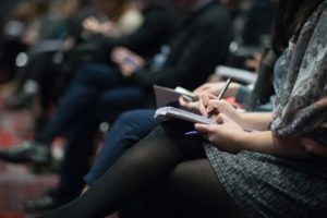 A row of people sat at an event. The woman in the foreground in writing in a notebook. 